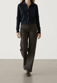 Granatowy dzianinowy kardigan z kołnierzem i guzikami, zestawiony z brązowymi spodniami typu wide-leg i ciemnymi butami na sznurówki, na neutralnym tle.