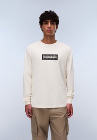 Langspeevige, crèmekleurige katoenen t-shirt met een gecentreerde zwart-witte logokader met tekst. Gecombineerd met lichtbruine cargobroeken.