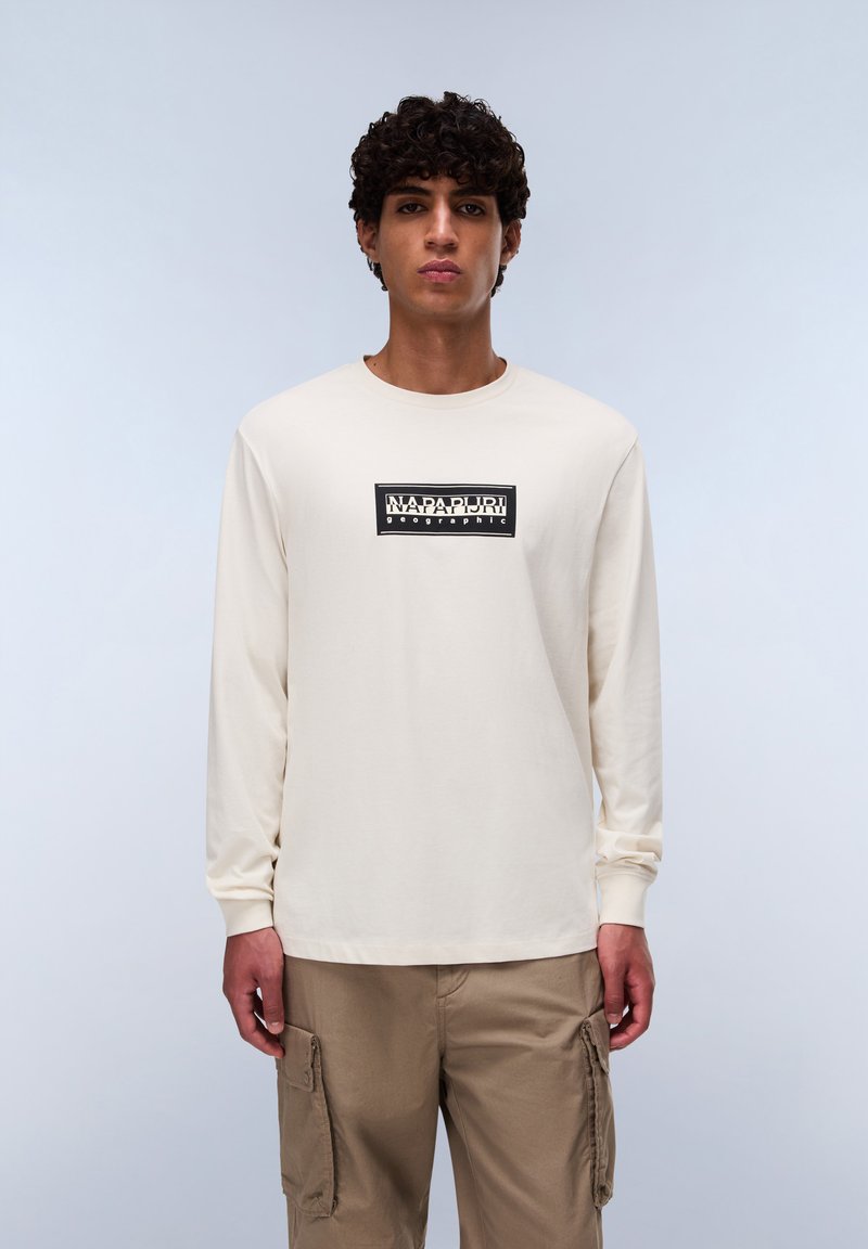 Langspeevige, crèmekleurige katoenen t-shirt met een gecentreerde zwart-witte logokader met tekst. Gecombineerd met lichtbruine cargobroeken.