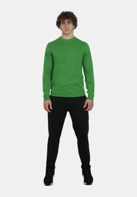 Maglione verde lavorato a maglia con scollo rotondo, polsini a coste e un piccolo logo. Abbinato a pantaloni neri slim-fit e scarpe nere.