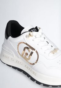 Sneaker bianco in ecopelle con accenti neri, logo in paillettes dorate, battistrada prominente e testo "MADE TO FEEL AMAZING" sulla suola.