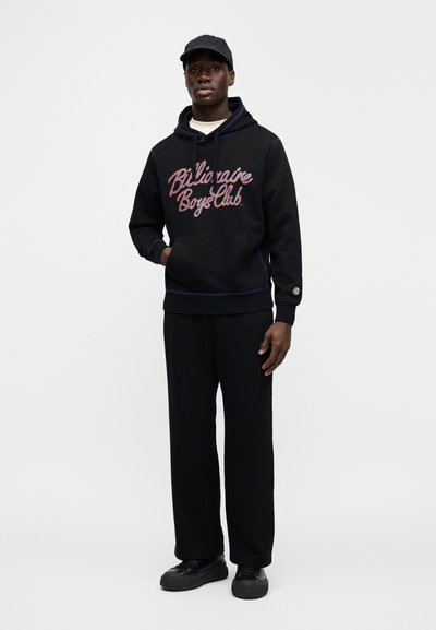 Μαύρο φούτερ με κουκούλα και κείμενο "Billionaire Boys Club", με κορδόνια και τσέπη καγκουρό. Συνδυασμένο με μαύρο παντελόνι φόρμας και αθλητικά παπούτσια.