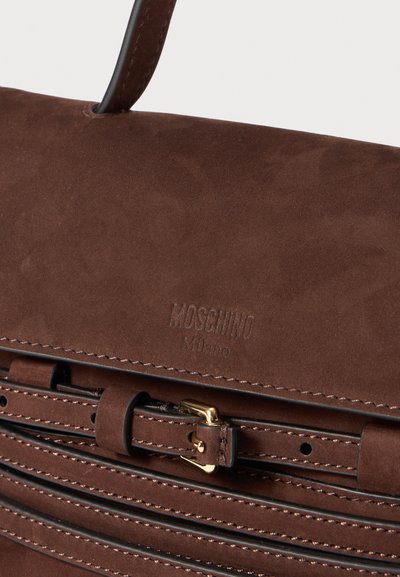 Bristu zamšādas rokassoma ar strukturētu dizainu, kurai ir zelta aksesuāri, sprādzes detaļa un iespiests logo "Moschino Milano."
