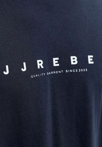 Σκούρο μπλε ύφασμα με λευκό κείμενο "JJREBE" και μικρότερο κείμενο κάτω που γράφει "ΠΟΙΟΤΙΚΟ ΕΝΔΥΜΑ ΑΠΟ ΤΟ 2023" σε ένα ρούχο.