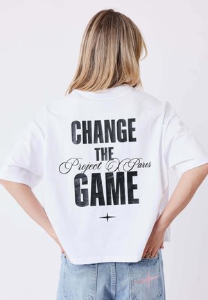 Femme aux cheveux blonds mi-longs porte un t-shirt blanc court avec le texte "CHANGE THE Project Pars GAME" et un jean bleu clair avec une étoile rose.
