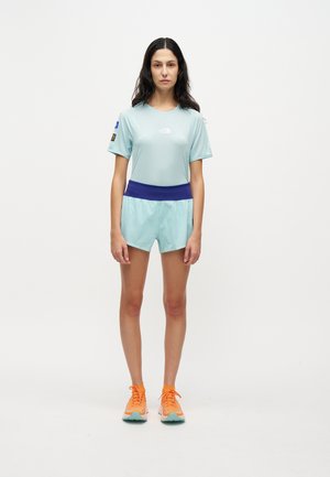 Donna in piedi che indossa una maglietta sportiva azzurro chiaro e pantaloncini con fascia in vita blu scuro e scarpe da corsa di arancione brillante su sfondo bianco.
