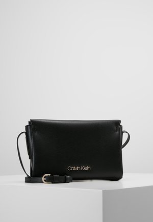 Czarna skórzana torebka Calvin Klein na ramię z cienkim regulowanym paskiem i złotym logo z przodu, prezentowana na białym piedestale.