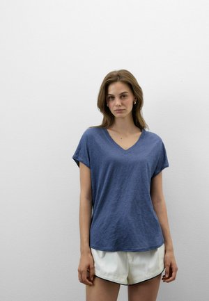 Młoda kobieta stoi przy gładkiej białej ścianie, ubrana w niebieską koszulkę z krótkim rękawem i dekoltem w serek oraz białe szorty z czarną lamówką.