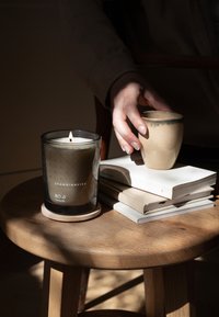Black glass candle with a wooden base, labeled "Skandinavisk RO Tranquility." Nearby, a textured beige ceramic cup rests on stacked books.