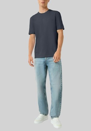 Mann trägt ein dunkelgraues Kurzarmhemd, hellblaue Jeans und weiße Turnschuhe, steht vor einem schlichten grauen Hintergrund.