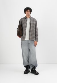 Geruit grijs en bruin shirt, witte T-shirt, losse lichtblauwe jeans en zwarte laarzen. Houdt een bruine leren tas vast.