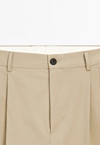 Beige trousers with a flat front, single black button closure, two front pockets, and a soft fabric texture. Neat stitching details visible.