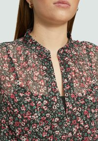 Florale Bluse mit schwarzem Hintergrund, die mit kleinen rosa und grünen Blumen verziert ist. Leichtes, semi-transparentes Material, Falten im Ausschnitt.