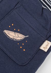 Navyblaues Stofffach mit einem bedruckten beigen Feder und orangefarbenen Punkten, befestigt an einem gestreiften Kleidungsstück mit einem beigen Etikett.