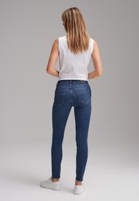 Femme debout, de dos, portant un haut blanc sans manches, un jean skinny bleu et des baskets blanches sur un fond gris uni.