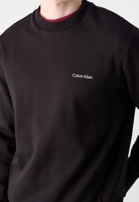 Sudadera negra hecha de una tela suave, con cuello redondo y un pequeño logo blanco de "Calvin Klein" en el lado izquierdo del pecho.