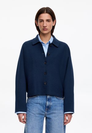 Kvinde iført en marineblå knappet cardigan over en lyseblå skjorte og lyseblå jeans, stående foran en ensfarvet hvid baggrund.