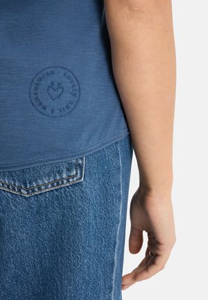 Person's arm beside blue denim jeans pocket and blue t-shirt with faint circular logo showing a heart and text around it.
