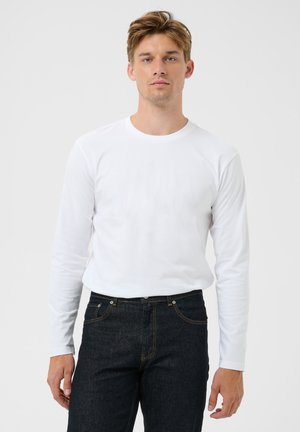 Hombre de pie vistiendo una camisa blanca lisa de manga larga y vaqueros azul oscuro frente a un fondo liso gris claro.