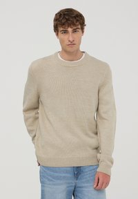 Beżowy sweter dzianinowy z okrągłym dekoltem i prążkowanymi mankietami. Luźny krój z teksturowanym wykończeniem, noszony z jasnoniebieskimi dżinsami.