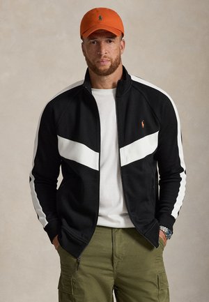 Homme portant une casquette orange, une veste noire zippée avec des bandes blanches, une chemise blanche, un pantalon cargo vert et une montre-bracelet, face à l'avant, les mains dans les poches.