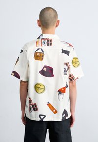 Camisa de manga corta hecha de tela ligera, con un fondo crema y gráficos coloridos de varios objetos y logotipos.