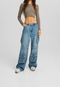 Pantalones cargo de mezclilla azul con piernas anchas, que cuentan con múltiples bolsillos y una textura desgastada. Combinados con un top marrón de manga larga y corto.