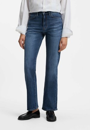 Kvinde iført blå bukser med lige ben, en hvid langærmet skjorte og sorte loafers står mod en ensfarvet baggrund.