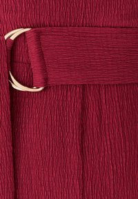 Burgundská textúrovaná látka s opaskom z umelej kože s kruhovou prackou v zlatej farbe, ktorá zdôrazňuje zložité detaily povrchu a dizajn.