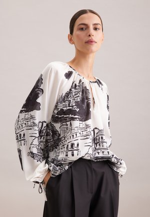 Blusa bianca con illustrazioni architettoniche nere, scollo arricciato e polsini elastici. Abbinata a pantaloni neri.