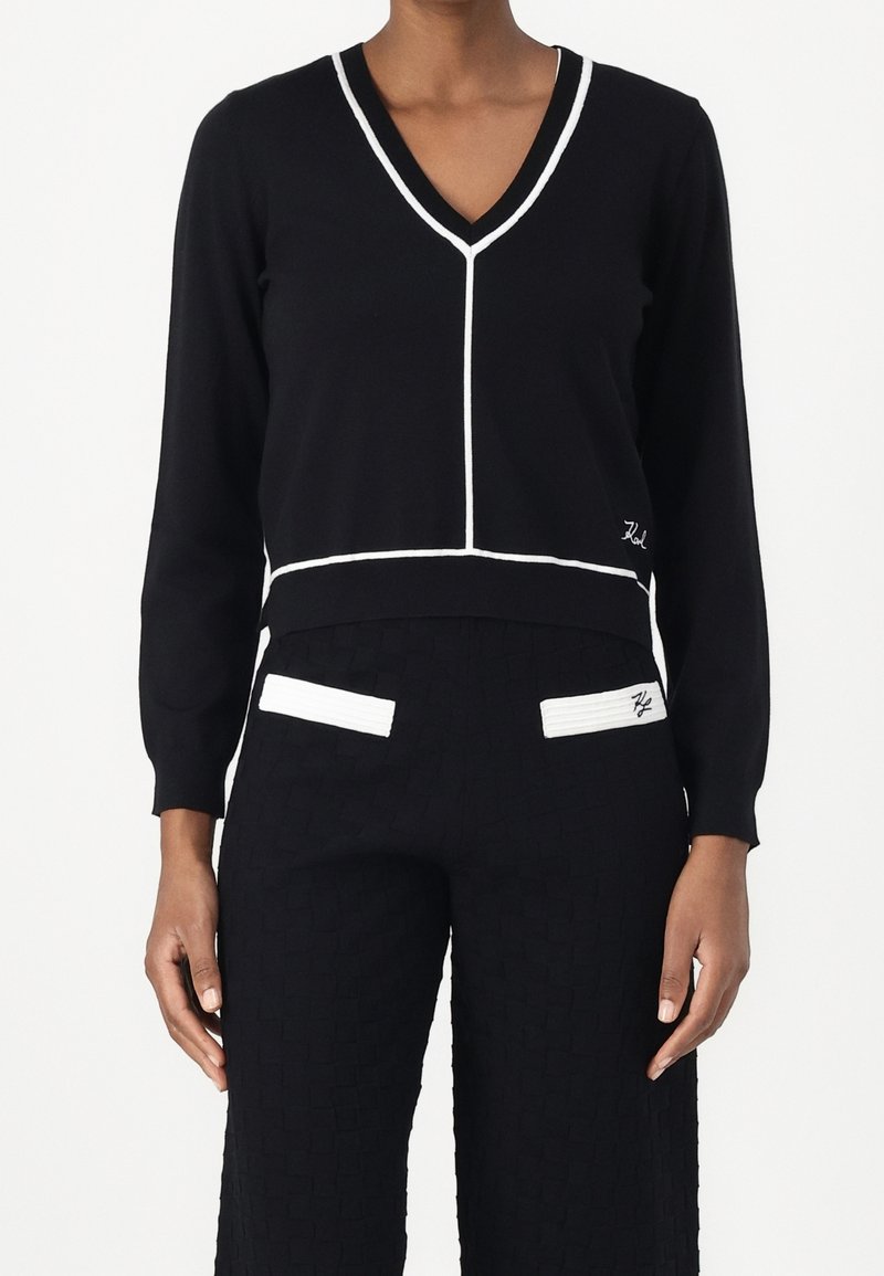 Pull noir à col en V avec bordure blanche et pantalon noir assorti avec des détails blancs sur les poches, porté par une personne debout.