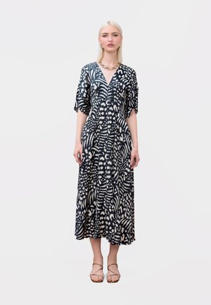 Mujer con cabello rubio vistiendo un vestido midi de escote en V con estampado animal en blanco y negro, y sandalias beige con tiras, de pie frente a un fondo simple.