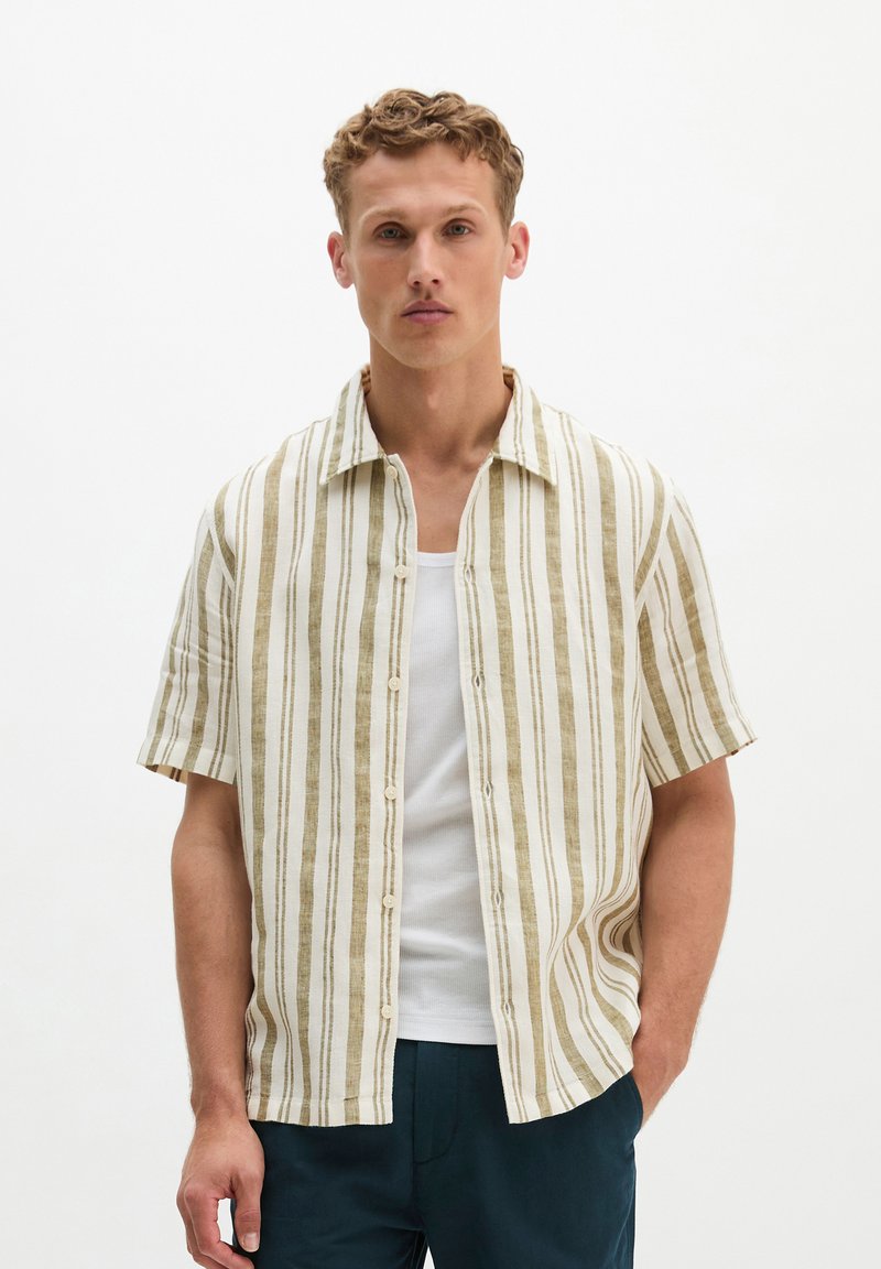 Giovane uomo con capelli corti e ricci che indossa una camicia beige e bianca a righe verticali a maniche corte sopra una canottiera bianca e pantaloni scuri.