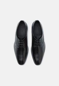 Zapatos de vestir de cuero negro con textura suave, diseño de punta afilada, cierre de cordones con seis agujeros y una suela ligeramente elevada. Marca visible en el interior.