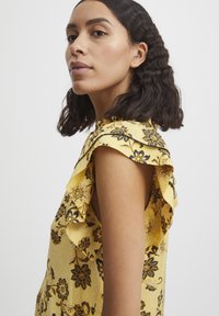 Blusa gialla a fiori con maniche a balze, motivi floreali neri e un tessuto a rilievo. La vista laterale mette in risalto i dettagli delle spalle e il taglio.