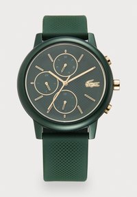 Grön klocka med en strukturerad rund urtavla, guldfärgade detaljer, tre små urtavlor och ett grönt strukturerat silikonarmband. Lacoste-logotypen finns på urtavlan.