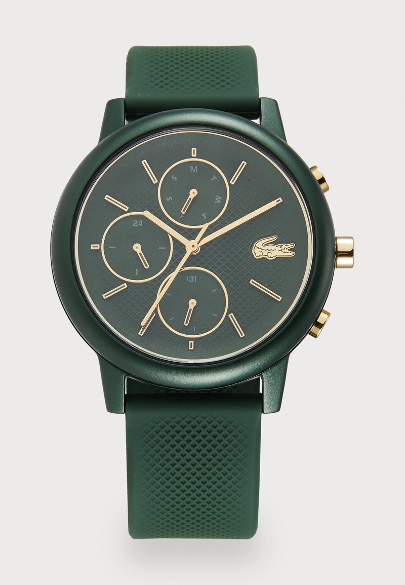 Grön klocka med en strukturerad rund urtavla, guldfärgade detaljer, tre små urtavlor och ett grönt strukturerat silikonarmband. Lacoste-logotypen finns på urtavlan.
