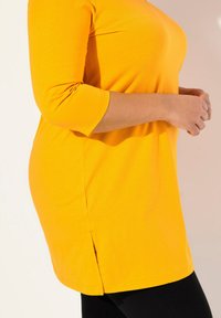 Gelbes, langes Tunika-Oberteil mit Dreiviertelärmeln. Verfügt über seitliche Schlitze und einen glatten, dehnbaren Stoff. Wird mit schwarzen Leggings getragen.