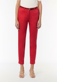Rote maßgeschneiderte Hose mit glatter Oberfläche, schmal zulaufendem Bein und einem braunen Gürtel mit rundem Verschluss. Trägt dazu nude-farbene Riemchenhighheels.