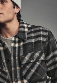 Camisa de cuadros para hombre hecha de felpa suave, con tonos gris, negro y blanco, con botones en el frente, dos bolsillos en el pecho y un cuello.