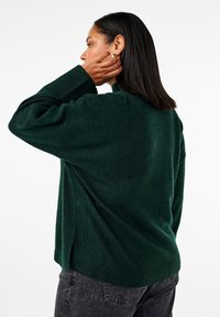 Ženska s srednje dolžino temnih las, oblečena v temno zeleno oversize pulover in črne kavbojke, se dotika svojih uhane, obrnjena levo na beli podlagi.