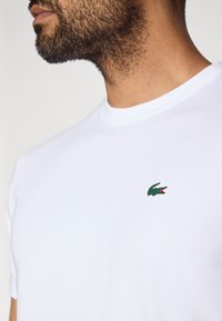 Λευκό βαμβακερό T-shirt με στρογγυλή λαιμόκοψη και κοντά μανίκια, με ένα μικρό πράσινο λογότυπο κροκόδειλου στο στήθος. Μαλακή υφή.