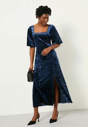 Navyblaues Samt-Maxikleid mit quadratischem Ausschnitt, kurzen Puffärmeln und einem Seitenschlitz; kombiniert mit einer schwarzen Handtasche und High Heels.