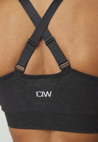 Svart sportsbra med kryssede stropper, teksturert stoff og hvit logo "ICIW" på den nedre kanten. Justerbare stroppere hardware synlige.