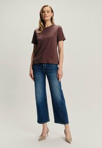 Brązowa koszulka krótki rękaw z okrągłym dekoltem, w połączeniu z szerokimi niebieskimi dżinsami i beżowymi butami na wysokim obcasie, stojąca na neutralnym tle.