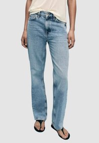 Personne portant un jean droit bleu clair, une chemise blanc cassé et des tongs noires, debout devant un fond uni et neutre.