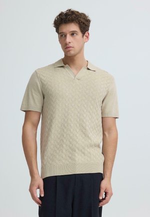 Młody mężczyzna o kręconych włosach, ubrany w beżową, teksturowaną koszulkę polo z krótkim rękawem i ciemne spodnie, spoglądający w bok na jednolitym tle.