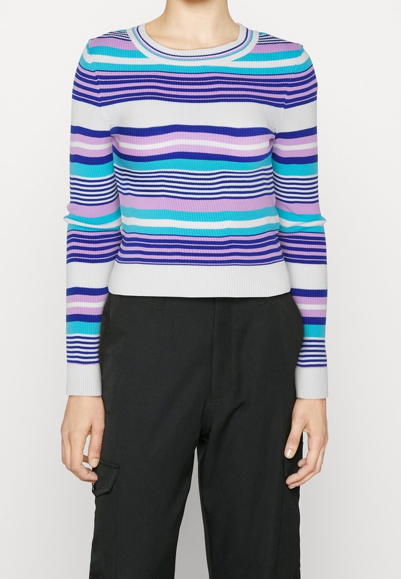 Personne portant un pull ajusté à manches longues avec des rayures horizontales en bleu, violet, rose, turquoise et blanc, associé à un pantalon noir.