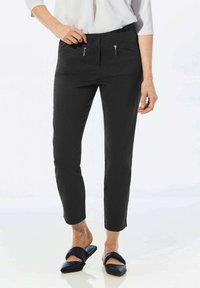 Pantalons noirs slim avec poches avant zippées, ceinture élastique et texture lisse, associés à des chaussures plates ouvertes.