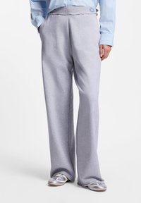 Άτομο που φοράει ανοιχτό γκρι παντελόνι φαρδιάς γραμμής, αθλητικά παπούτσια σε μεταλλικό ασημί και λευκό και ένα μπλε ριγέ πουκάμισο με κουμπιά, με τα χέρια στις τσέπες.